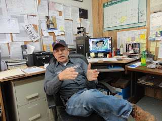 A man in work clothes and a cap sits in an office chair, gesturing with his hands. Behind him are cluttered desks, a computer showing a racing scene, and bulletin boards filled with papers—suggesting plans for a super-capacity grease interceptor.