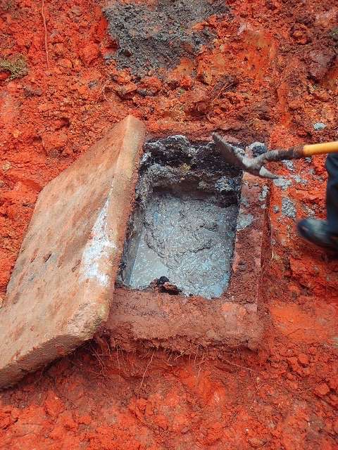 A person uses a shovel to open a rectangular concrete super-capacity grease interceptor partially filled with water, embedded in reddish soil. The concrete lid is propped to the side.