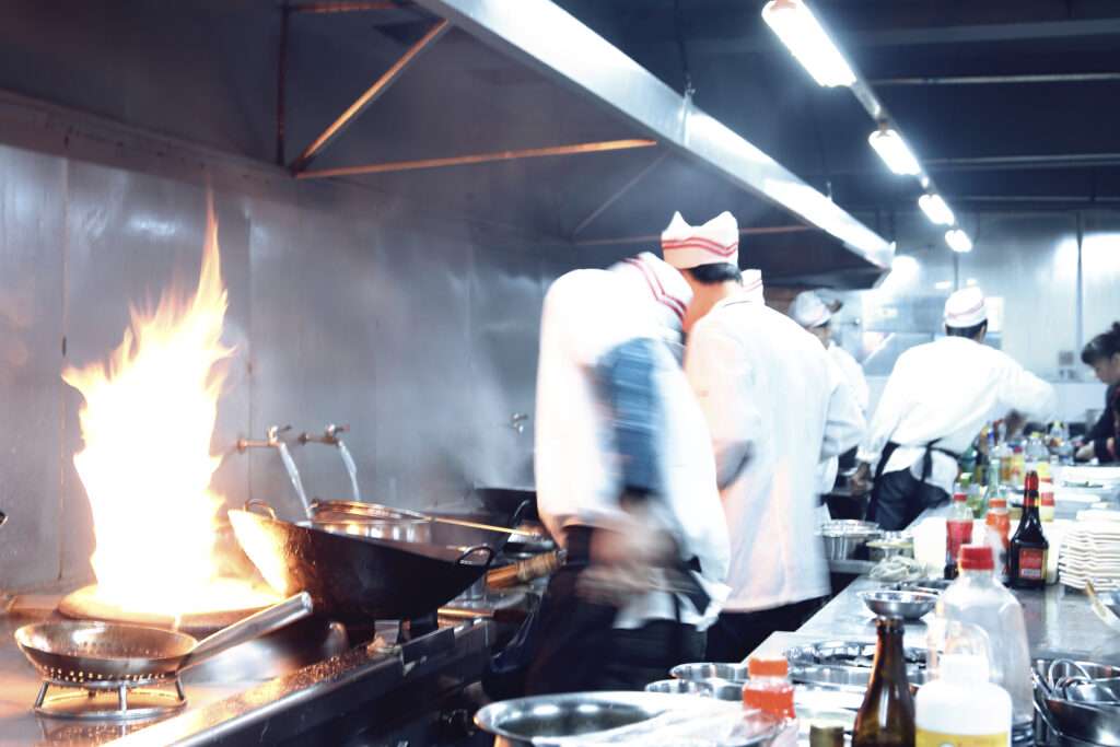 A busy commercial kitchen with chefs in white uniforms and hats preparing food. Flames rise from a pan, while ingredients are spread across the counters—powered by a super-capacity grease interceptor for restaurants to ensure smooth operation.