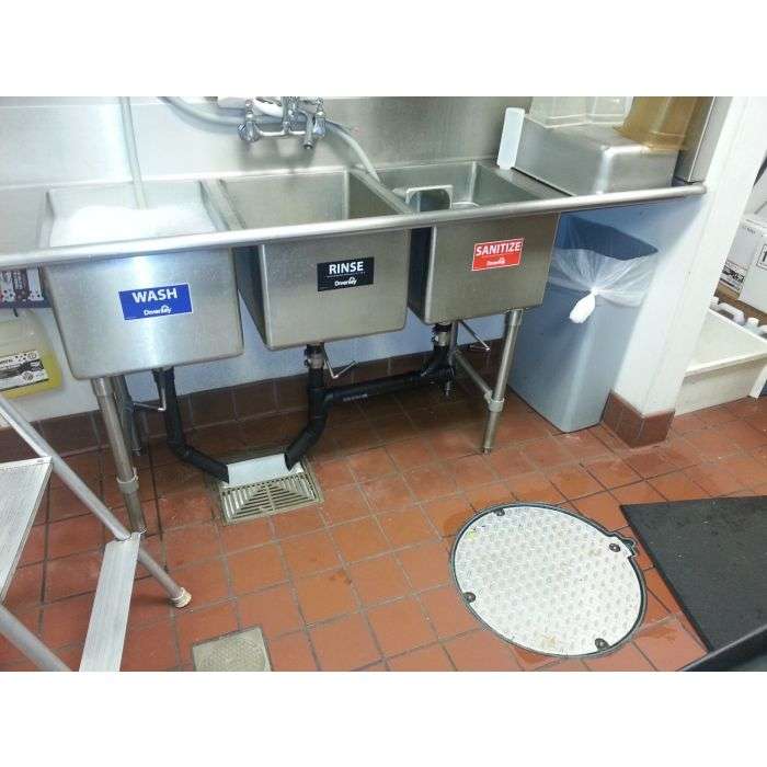 A commercial kitchen features a three-compartment sink marked “WASH,” “RINSE,” and “SANITIZE,” with a faucet above, a TZ-600 Trapzilla Grease Interceptor below, a trash can to the right, and tiled flooring with a drain and round access cover.